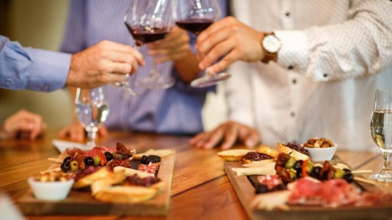 Foto de una mesa con tapas de comida y dos manos con sendas copas de vino, brindando.